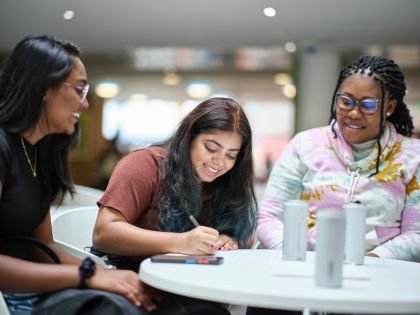 Image shows three female students sitting around a table with the one in the middle writing