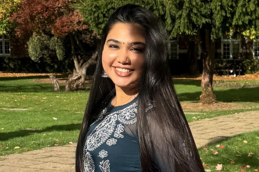 Smiling woman with long dark hair standing in a sunny park with trees and grass in the background.